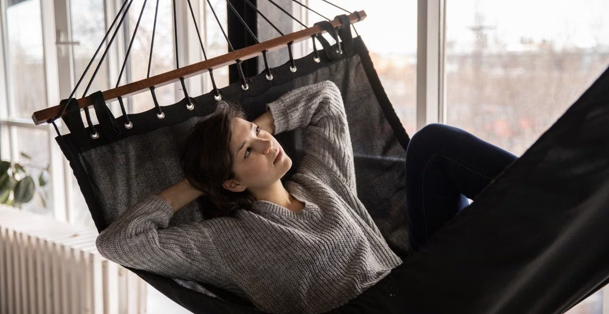 Thoughtful female laying with hands overhead in hammock with black canvas inside of apartment and looking away