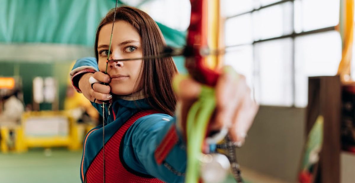 Woman archer aiming with bow indoors, emphasizing precision and focus.
